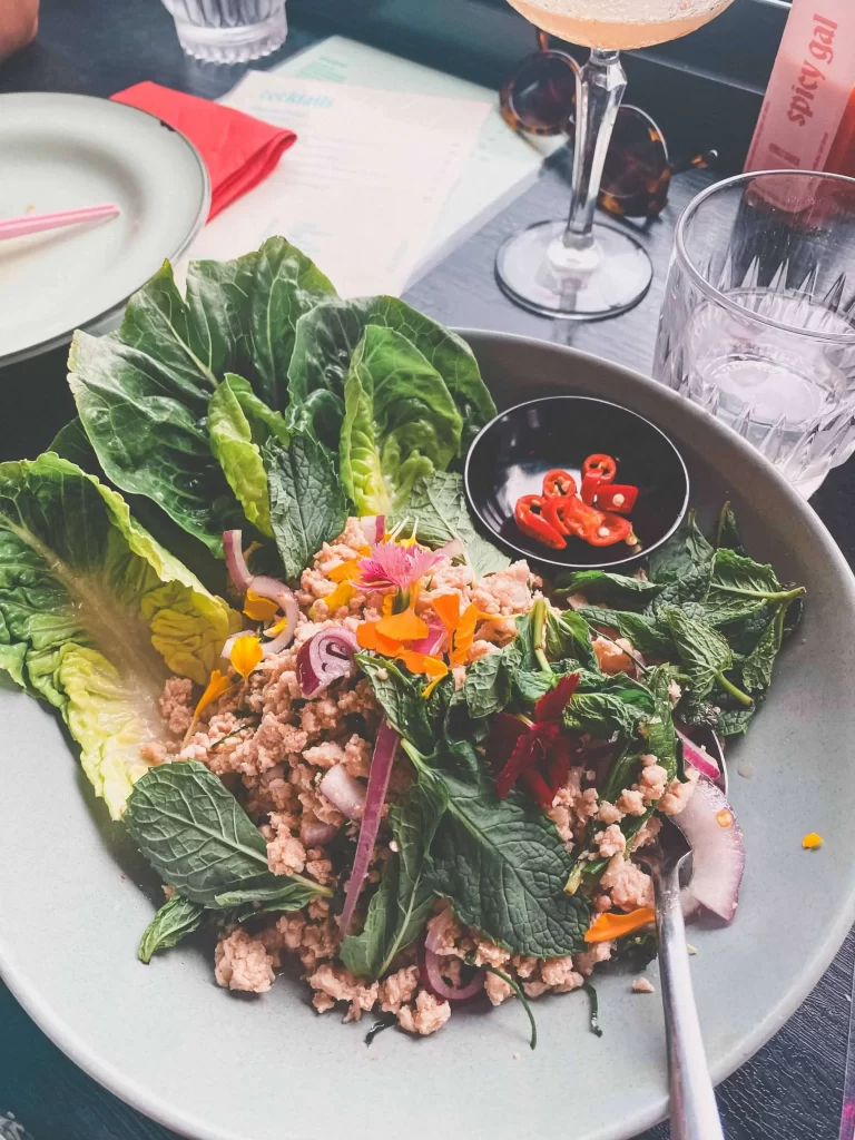 A plate of Thai larb salad with minced meat, fresh lettuce leaves, herbs, sliced red onions, and a small bowl of sliced red chili peppers on the side, beautifully presented at Maew Maew Terrigal. Drinks and utensils are visible in the background.