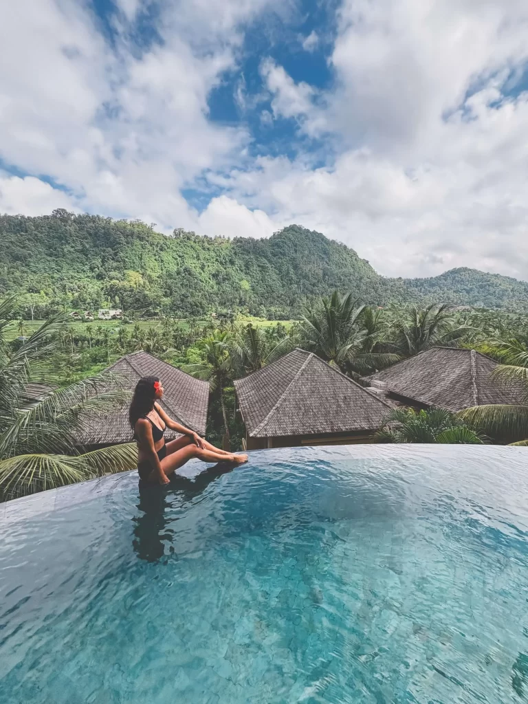A person in a swimsuit sits by the edge of an infinity pool at Wapa di Ume Sidemen, overlooking lush green hills, palm trees, and rooftops under a partly cloudy sky.