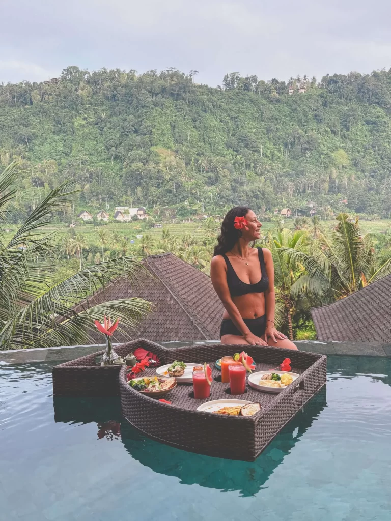A woman in a bikini with a red flower in her hair sits on the edge of a pool with a floating breakfast tray at Wapa di Ume Sidemen. Lush green hills and rooftops are visible in the background.