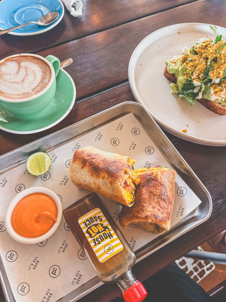 A tray with a burrito cut in half, a lime wedge, a small bowl of orange sauce, and a bottle of hot sauceβjust the kind of spread youβd expect at Milk and Madu. Nearby, thereβs a green cup of coffee and a plate with avocado toast topped with herbs.