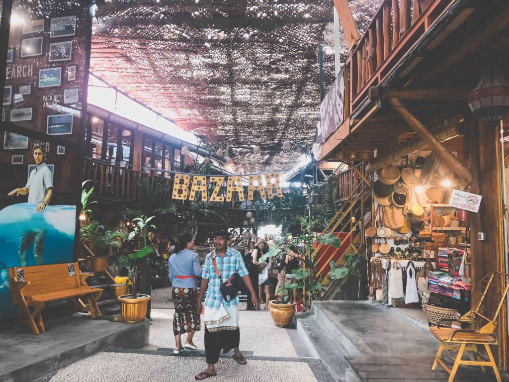 Indoor bazaar with wooden decor, hanging sign reading BAZAAR, people browsing shops, hats, bags, and souvenirs displayed—sunlight filters through a patterned ceiling and green plants surround the entrance area. This vibrant scene captures the essence of Love Anchor Market, making it one of the top things to do in Canggu.