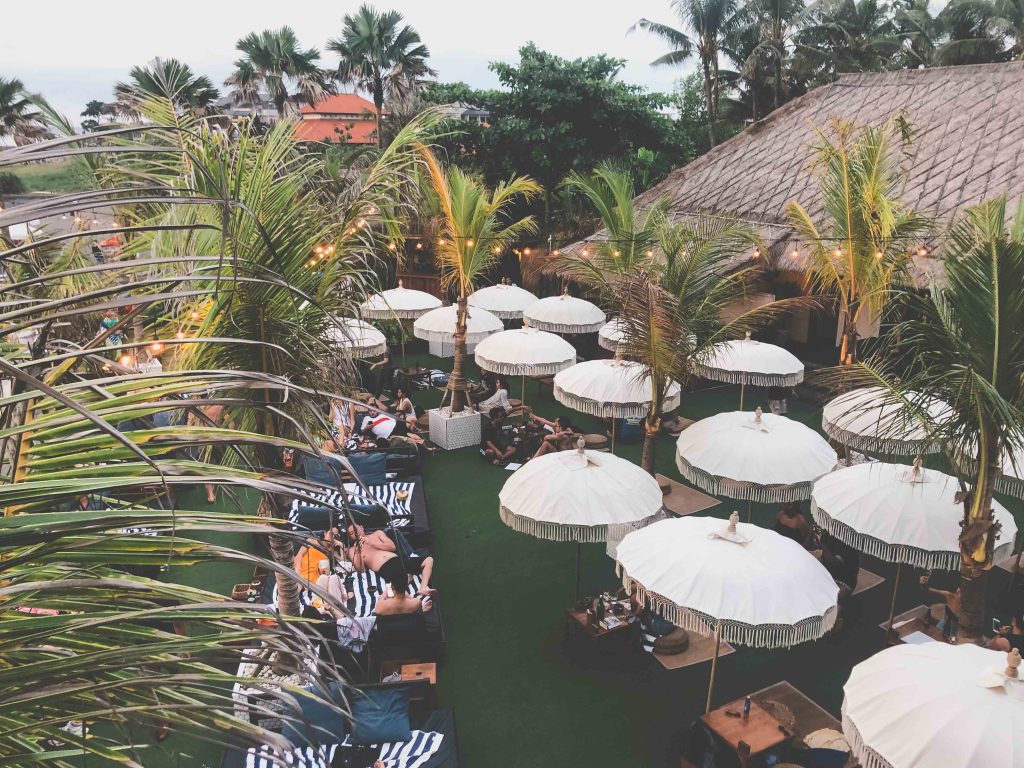 Outdoor lounge at The Lawn Canggu with white umbrellas, palm trees, people relaxing on striped chairs, and a thatched-roof building, creating a tropical, laid-back atmosphere.