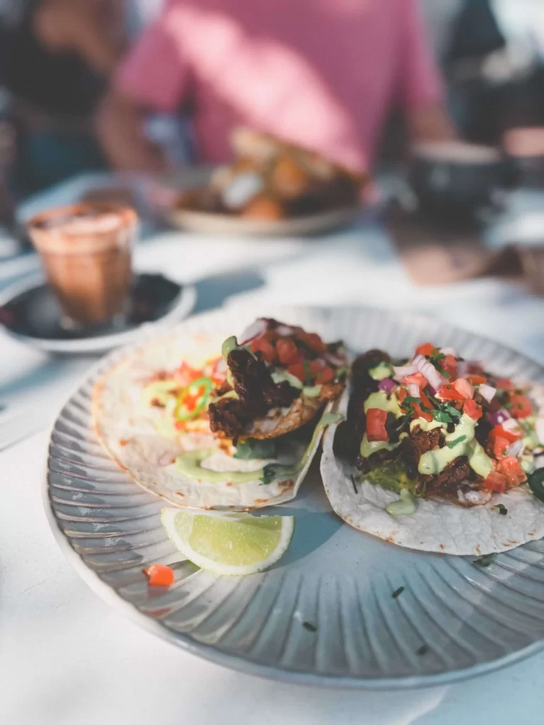 Two tacos topped with diced tomatoes, greens, and sauce are on a plate with lime wedges at Lune Cafe Central Coast. A blurred person and drink are in the background.