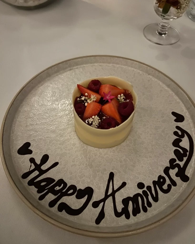 A white dessert topped with strawberries and raspberries sits on a plate with Happy Anniversary written in chocolate along the rim.