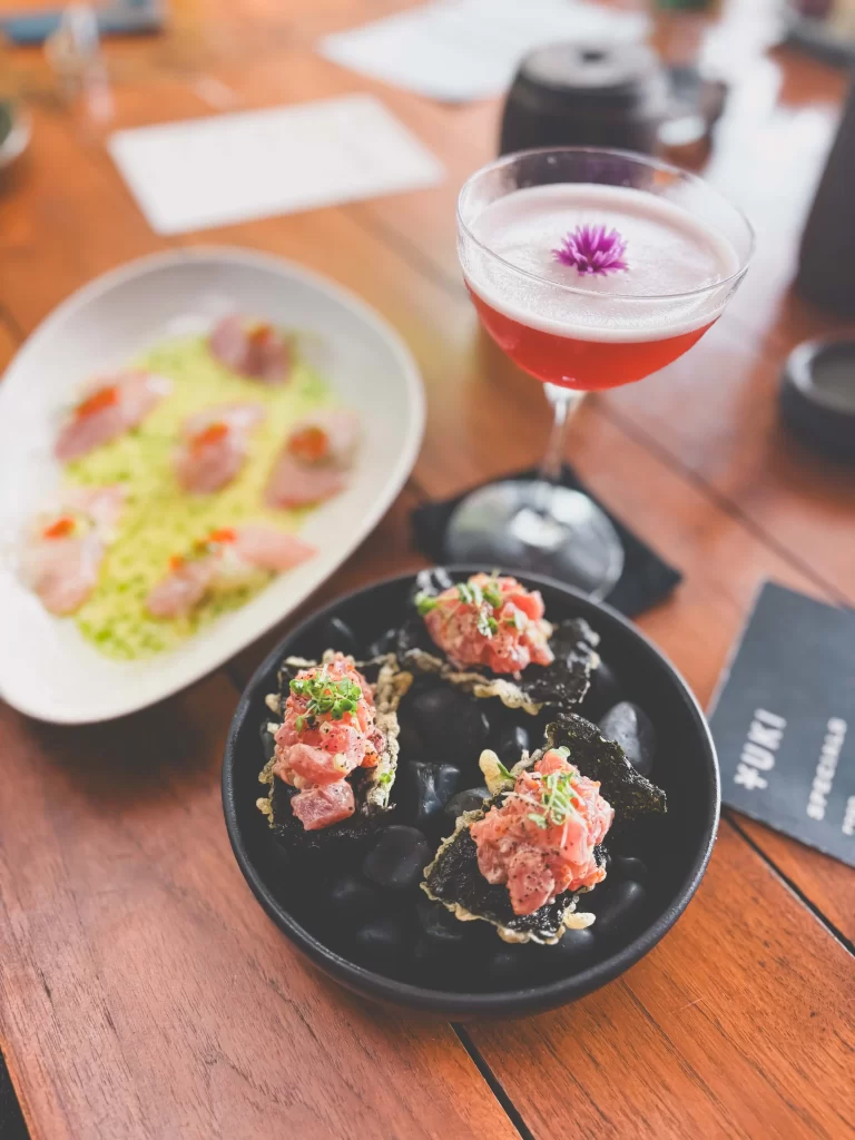 A cocktail with a purple flower garnish sits beside a bowl of appetizers topped with diced fish and herbs, and a plate of sliced fish on a bed of green garnish, all on a wooden table at Yuki, best restaurant Uluwatu.