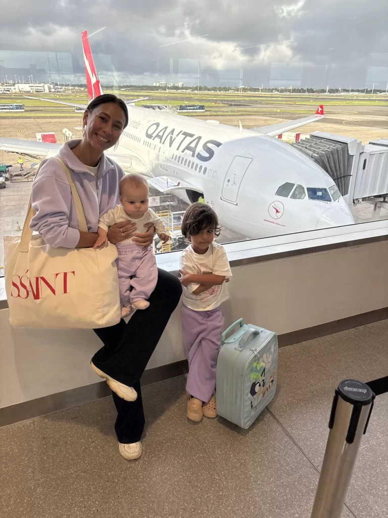 A woman holding a baby stands next to a young child with a suitcase in an airport terminal, possibly ready to discover the Bali trip cost as a Qantas airplane waits outside the window behind them.