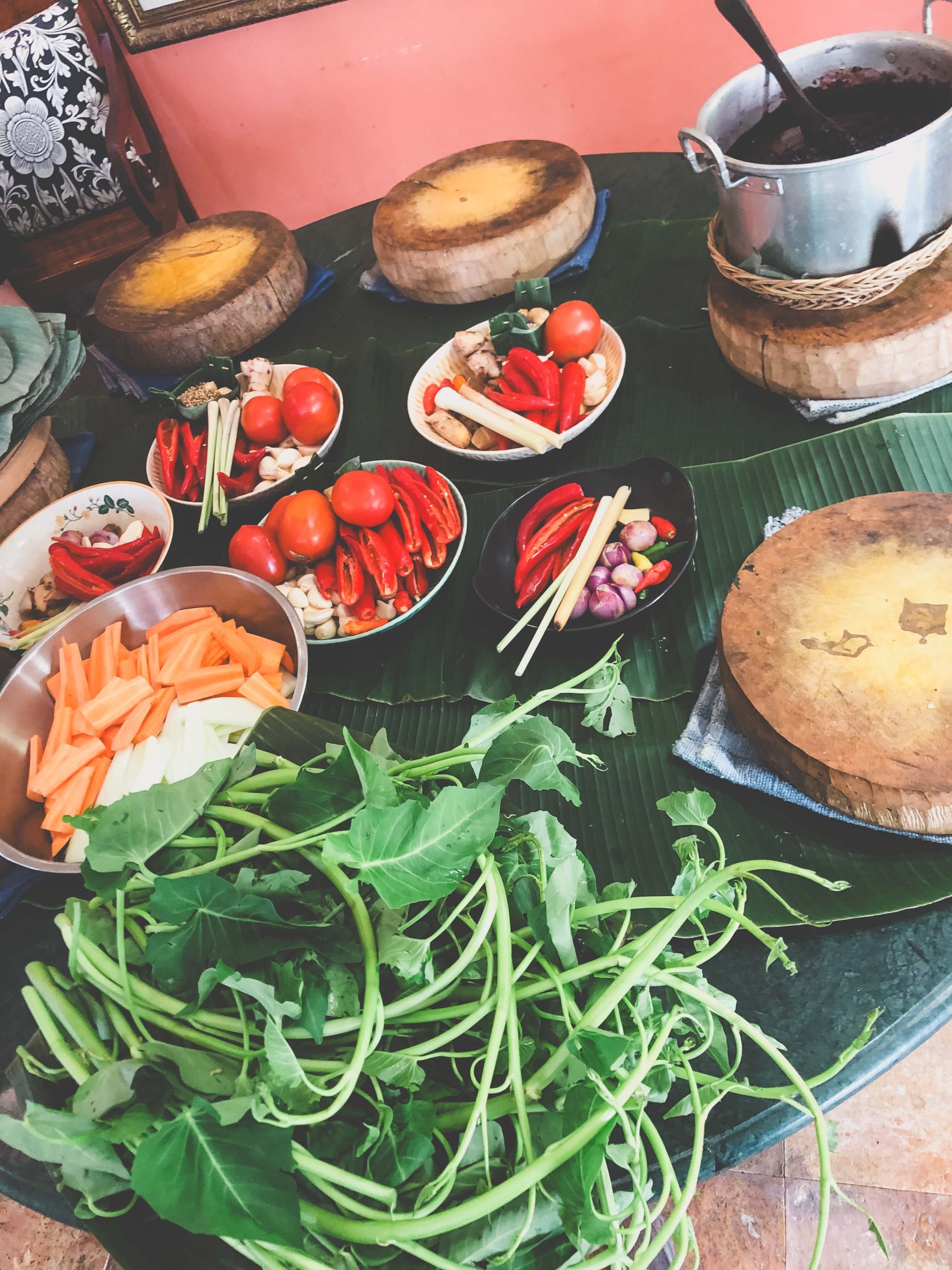 Various fresh vegetables, including leafy greens, tomatoes, carrots, and chili peppers, are arranged in bowls on a round table covered with banana leaves—a perfect sight for those exploring cultural Things to Do in Bali with Kids. A large pot sits on a wooden stand in the background.