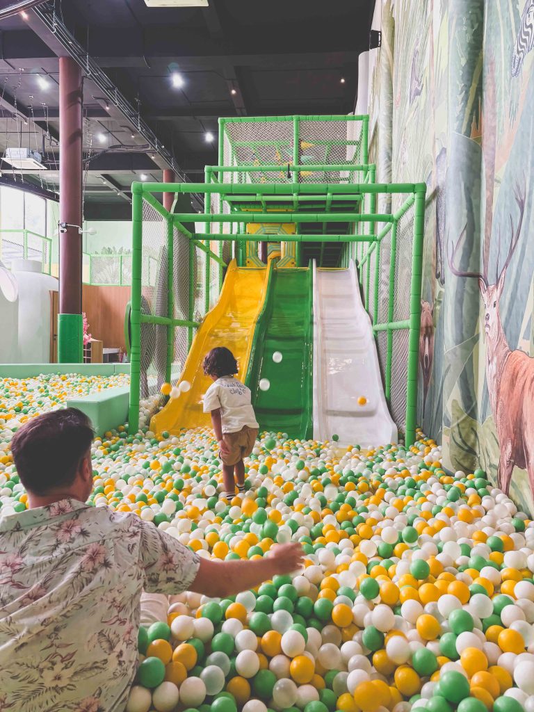 A child runs towards green and yellow slides in the Mai Main Playground, filled with yellow, green, and white plastic balls, while an adult sits nearby. Murals of animals and trees decorate the walls.