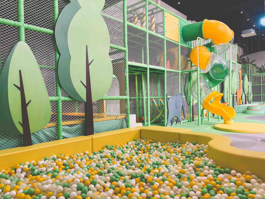 Mai Main Playground features a clean, empty indoor play area with a yellow, green, and white ball pit, green tree decorations, climbing structures, and a tall yellow and green tube slide in the background.