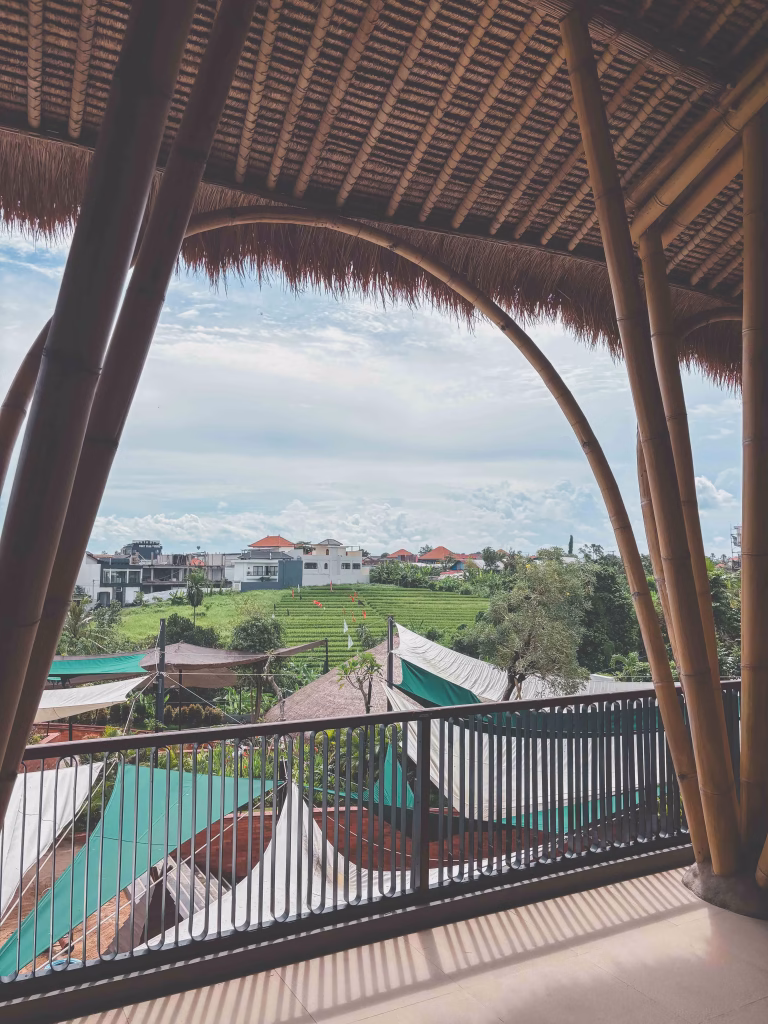 View from under a bamboo roof, looking out over lush green rice paddies in Canggu, houses with red roofs, and tent-like market stalls, with a partly cloudy sky in the background.