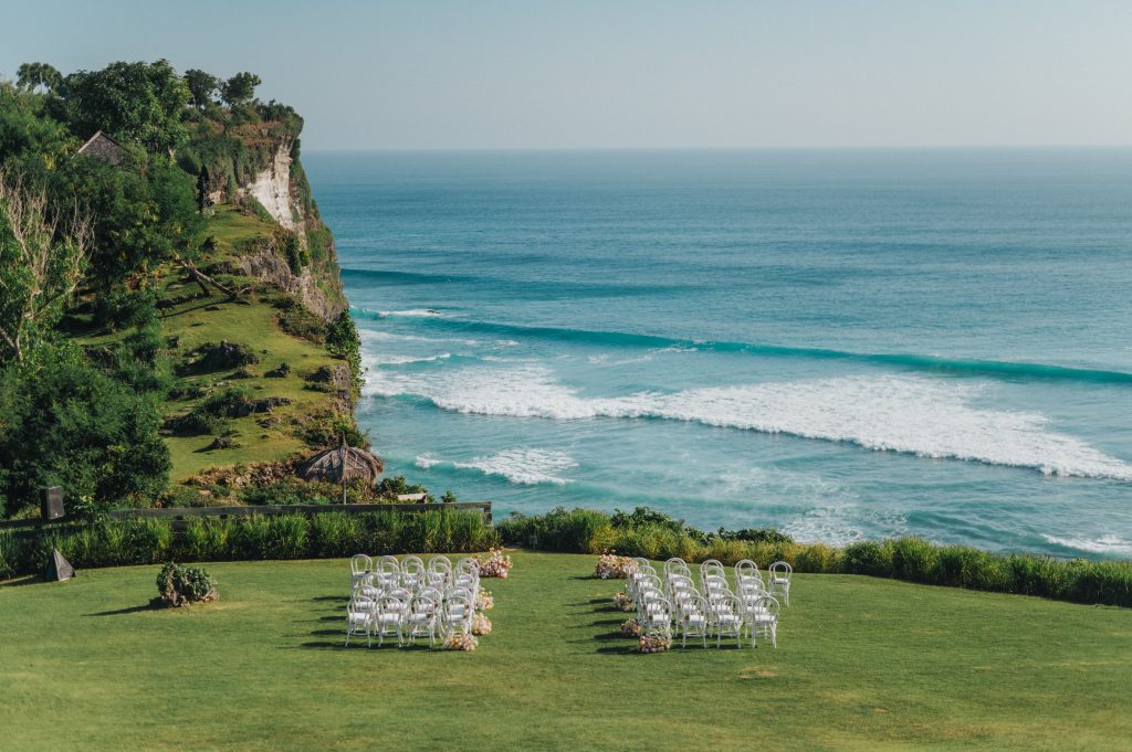 Outdoor wedding ceremony setup on a green cliffside lawn overlooking the ocean with rows of white chairs facing the water. Waves roll below the rocky cliff and tropical greenery surrounds the ceremony area. The image highlights a scenic seaside wedding location with dramatic coastal views.