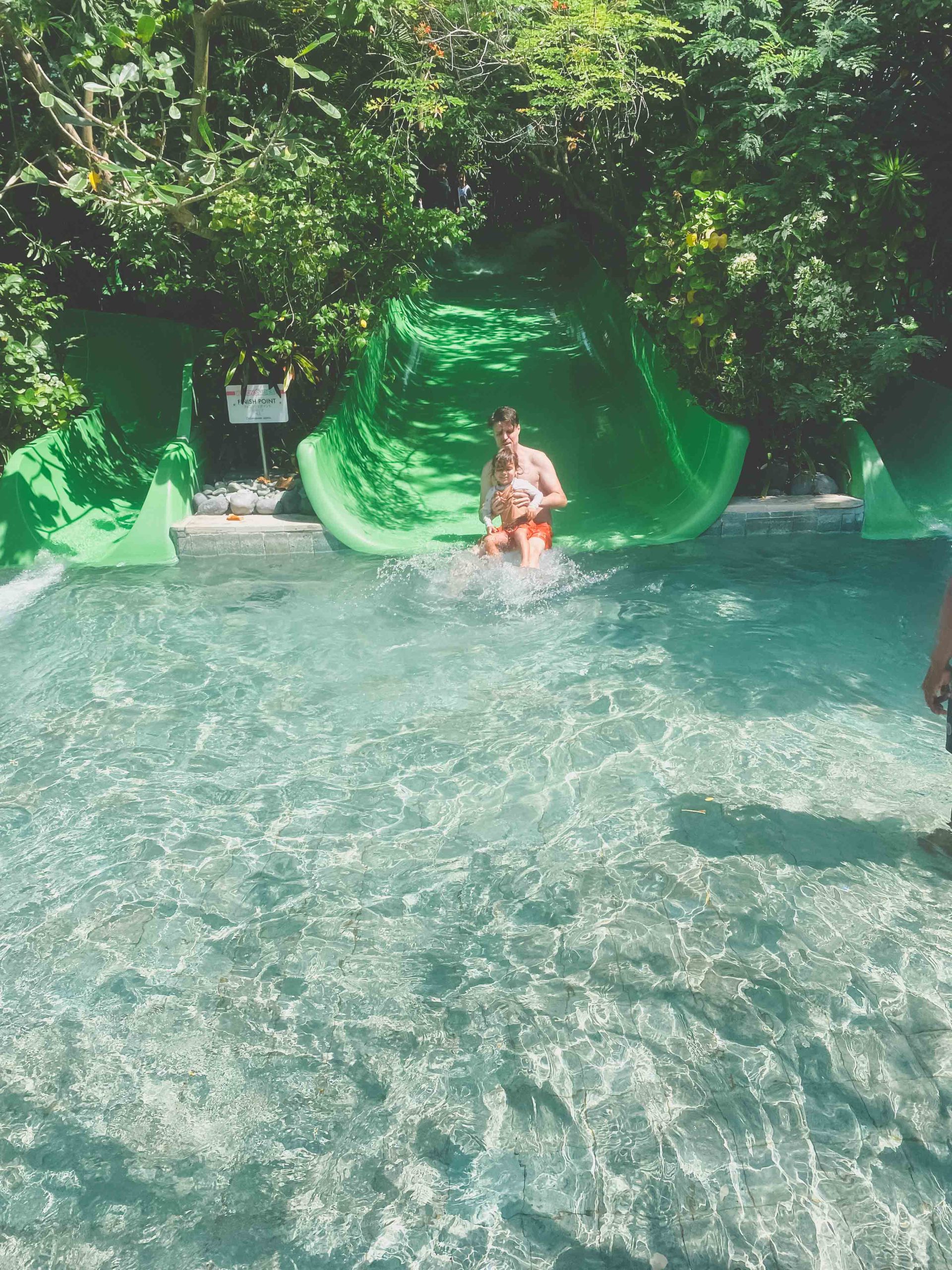 Two people come down a bright green water slide together, splashing into a clear blue pool surrounded by lush green plants and trees—one of the top things to do in Bali with kids for endless tropical fun.