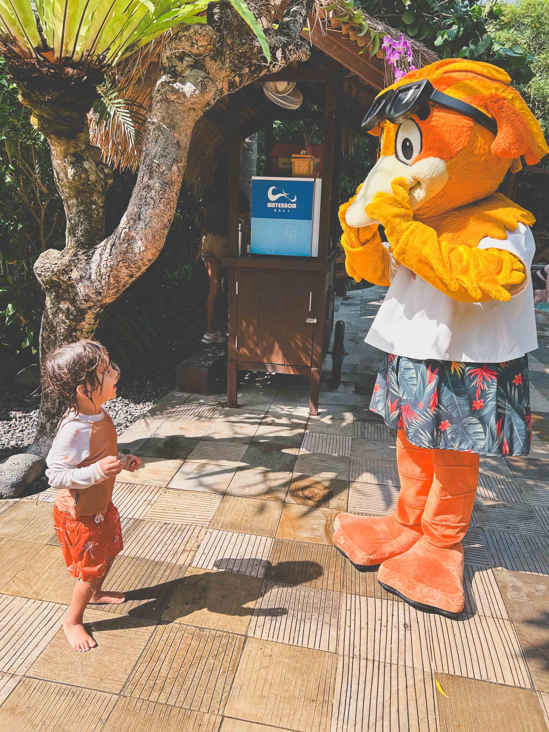 A young child in wet clothes stands on a tiled path outdoors, gazing up at a large orange animal mascot in sunglasses and floral shorts. Lush greenery surrounds them—a perfect moment among the many Things to Do in Bali with Kids.