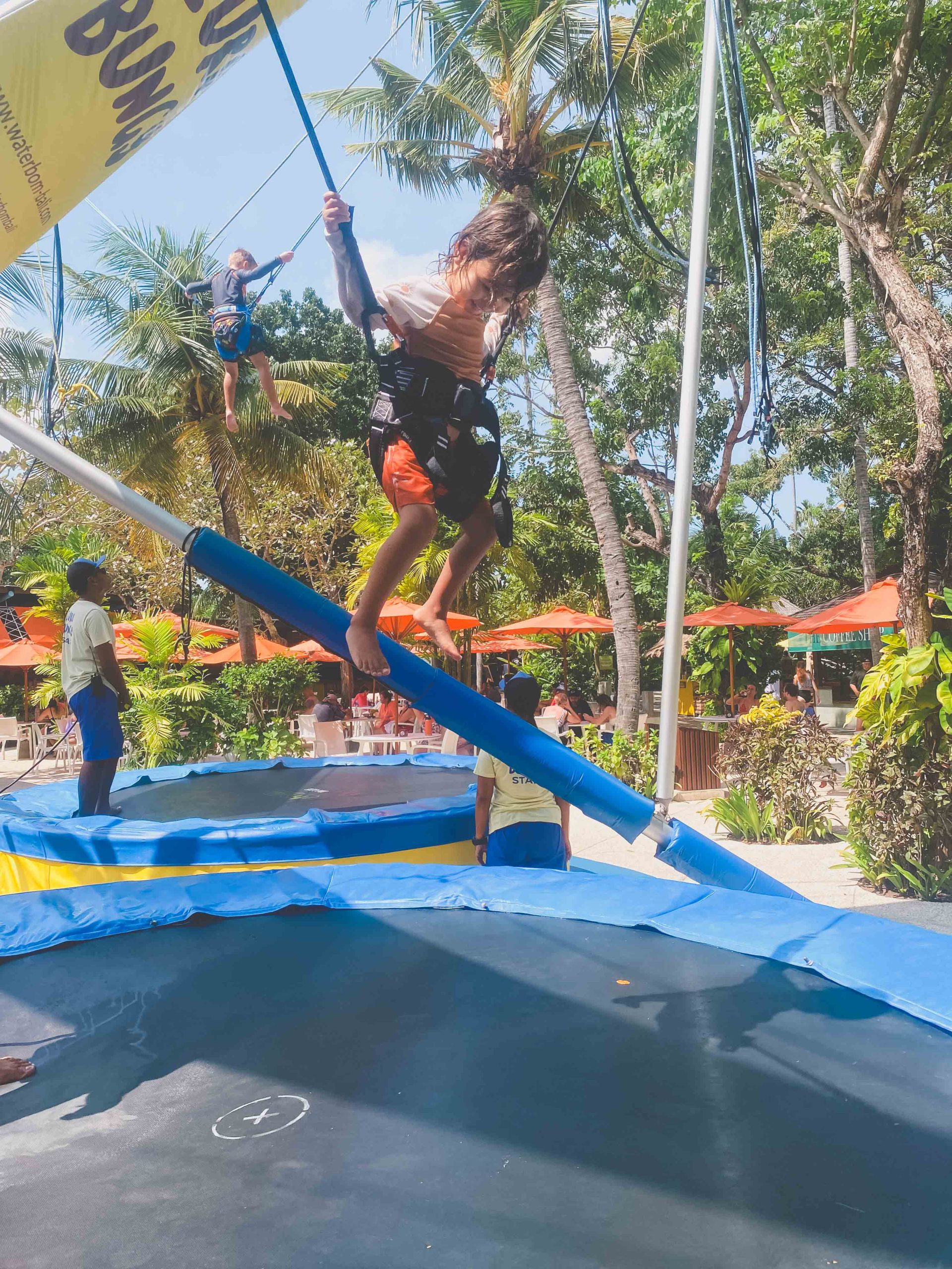 A child in a harness bounces on a trampoline with a bungee cord setup outdoors, surrounded by lush trees and orange umbrellas. Another child jumps high in the background—one of the top things to do in Bali with kids for extra fun.