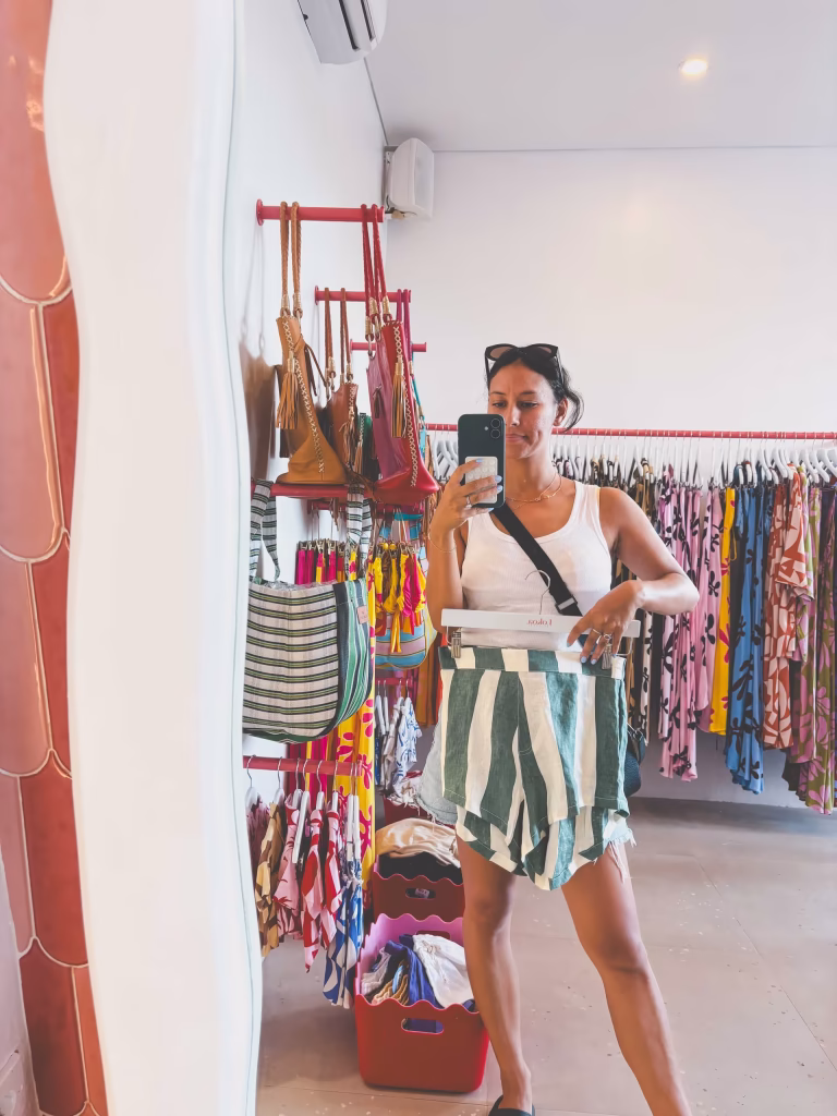 A woman takes a mirror selfie in a clothing store, holding green and white striped shorts. She wears a white tank top, sunglasses on her head, and a crossbody bag. Colorful clothes and handbags hang on racks behind her.