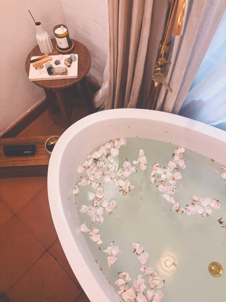 A white bathtub filled with water and floating rose petals sits beside a small wooden table holding a candle, white vase, stones, and incense, creating an atmosphere of serene alam wellness next to beige curtains.