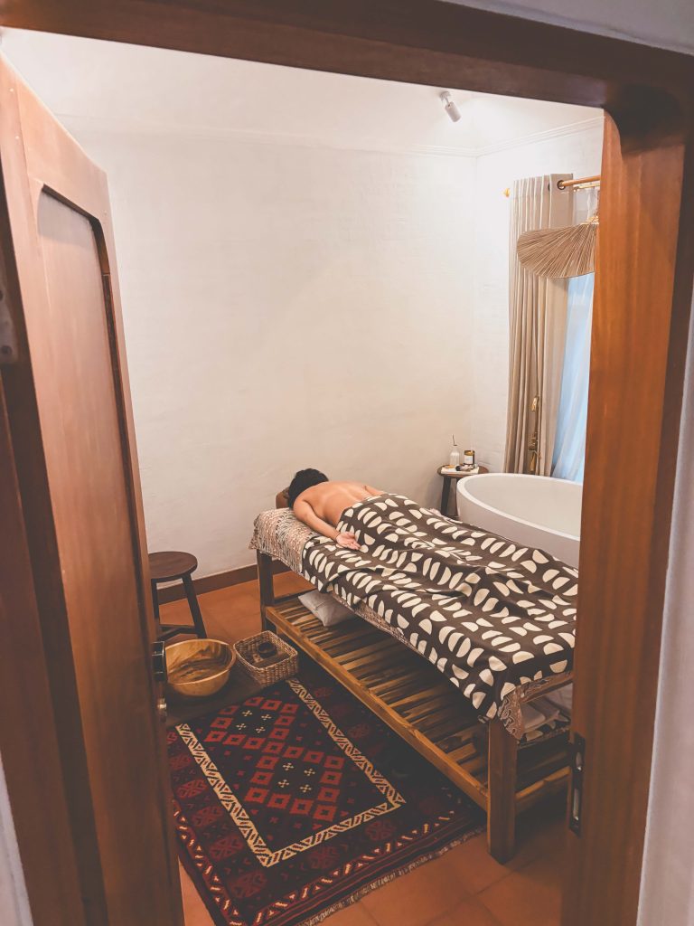 A person lies face down on a massage table covered with a patterned blanket in a cozy, softly lit spa room near Seseh Beach, with a rug, wooden stool, and a bathtub by the window.