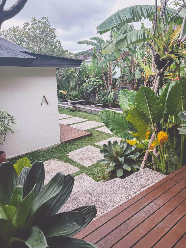 A tropical garden with large green plants, banana trees, and a wooden deck. Stone pathways lead past a white building with the letter A on the wall, under a cloudy sky.