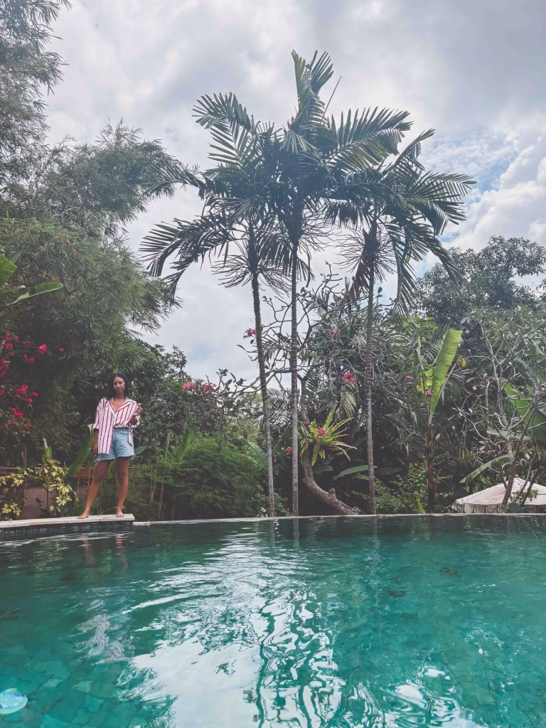 A woman in a red and white striped shirt stands at the edge of a clear turquoise pool, surrounded by lush tropical trees and plants under a cloudy sky.