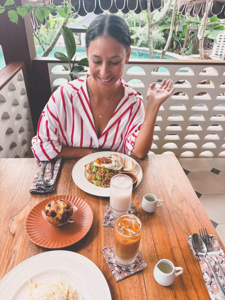 A woman in a red-and-white striped shirt sits at a wooden table with various foods and drinks, including noodles, iced coffee, a muffin, and milk, smiling and looking down at her meal in a tropical outdoor setting.