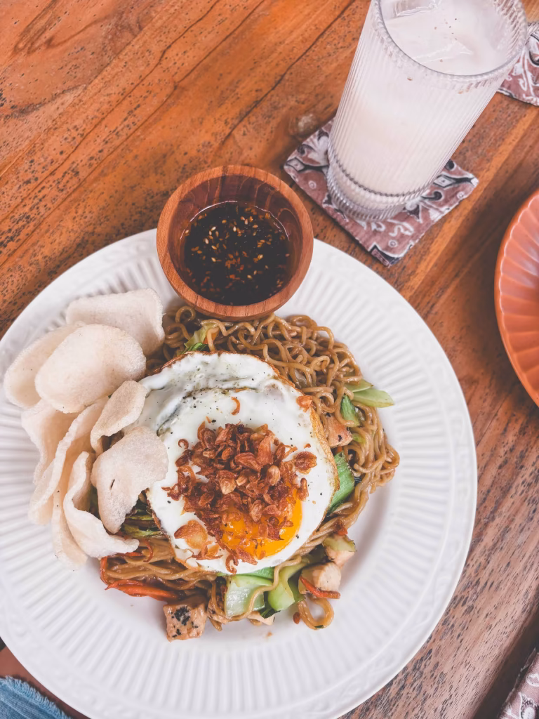 A plate of stir-fried noodles with vegetables, topped with a sunny-side-up egg and crispy shallots, served with prawn crackers and a small bowl of dipping sauce, next to a glass of iced drink on a wooden table.
