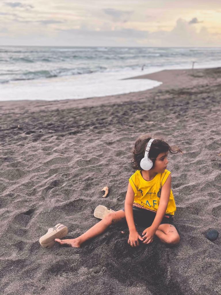 A young child wearing white headphones and a yellow shirt sits on dark sandy beach, looking to the side. One shoe is off and the ocean waves are visible in the background under a cloudy sky.