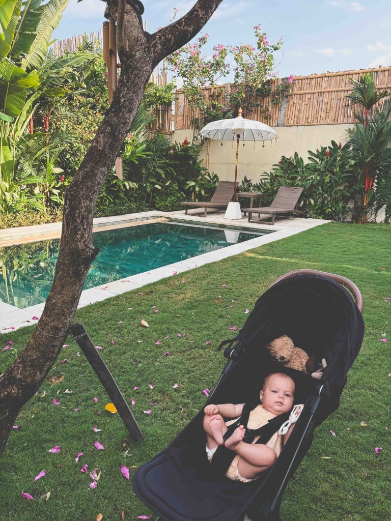 A baby in a stroller sits on a grassy lawn near a pool, just moments from Seseh Beach. Tropical plants, bamboo fencing, lounge chairs, and a white umbrella surround the pool while pink flower petals are scattered on the grass.