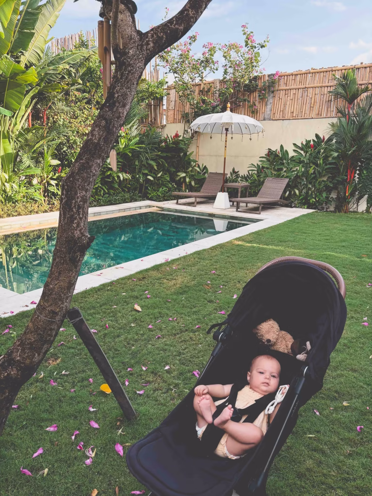 A baby lies in a stroller on the grass near a swimming pool. The pool is surrounded by lounge chairs, an umbrella, and lush green plants and trees. Flower petals are scattered on the grass.
