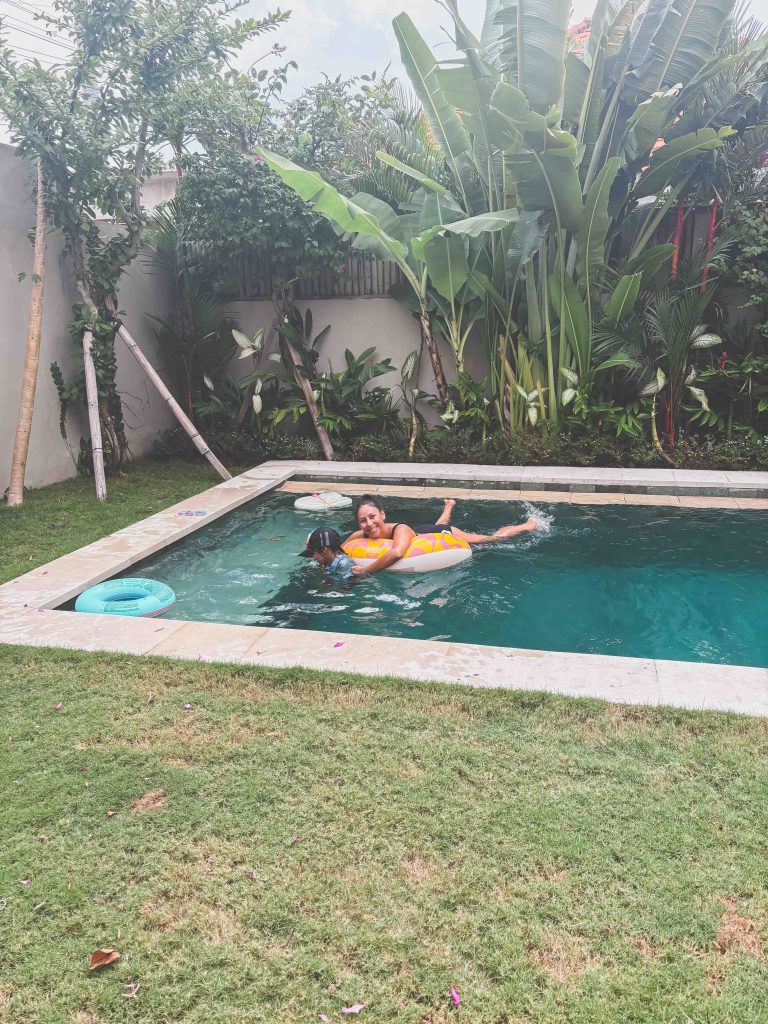 A woman in a yellow swimsuit floats on a pool ring in a small backyard pool surrounded by lush greenery, evoking the tropical vibes of Seseh Beach, with grass covering the area around the pool.