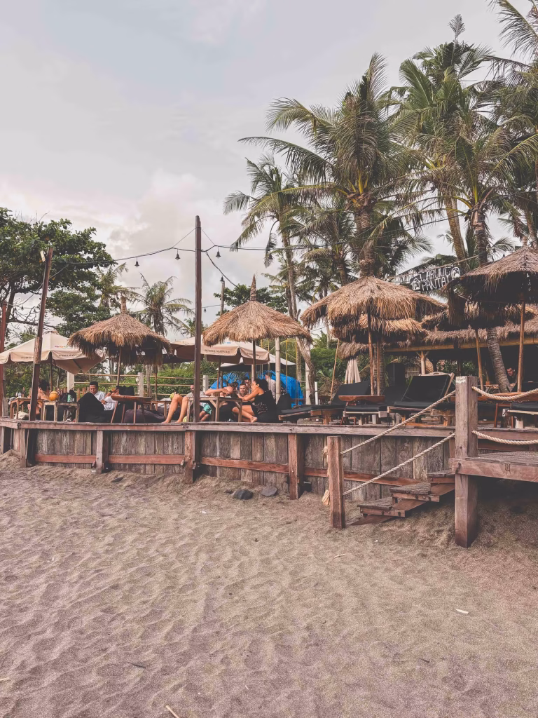 A beachside bar with wooden railings, thatched-roof umbrellas, lounge chairs, and people relaxing under palm trees; string lights are hung above the seating area, and the sandy beach is in the foreground.
