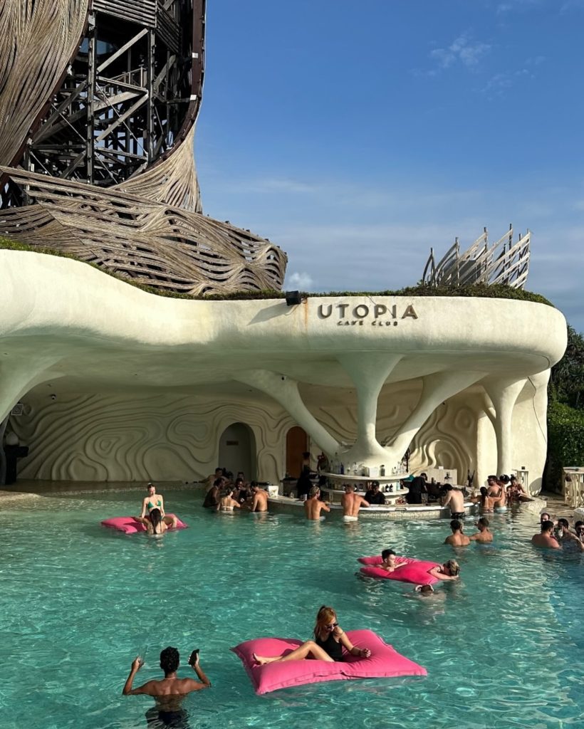 People relax and socialize in a pool at Utopia Café near Seseh Beach, some sitting on pink floats, while others gather at the swim-up bar beneath a sculpted, white, futuristic structure. The sky is clear and sunny.
