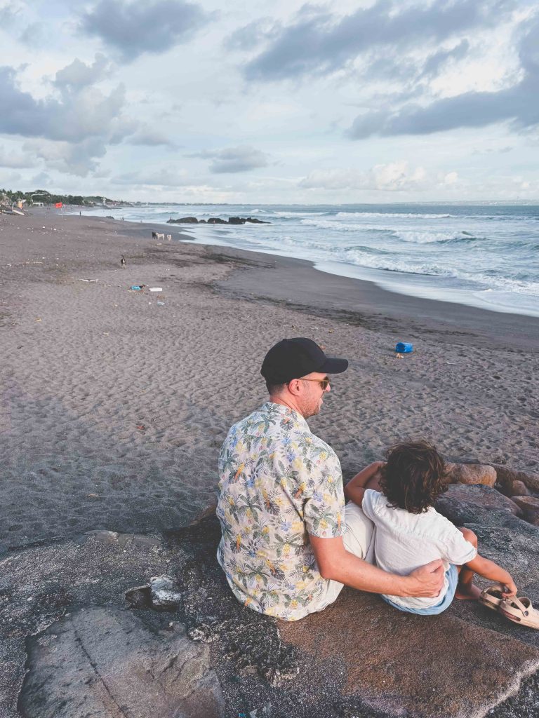 A man in a floral shirt and cap sits on a rock at Seseh Beach, embracing a young child. They face the ocean, with waves rolling onto the sandy shore and a cloudy sky in the background.