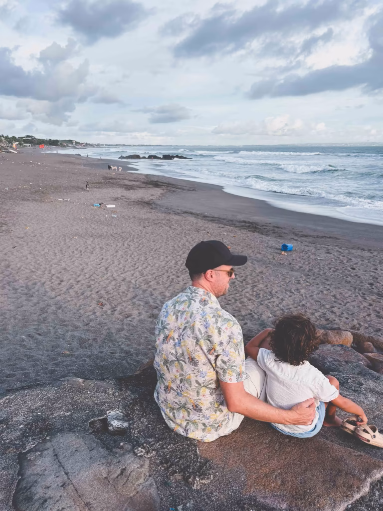 A man in a floral shirt and black cap sits on a rock at the beach, holding a child. They look out at the ocean waves under a cloudy sky, with a sandy shoreline stretching into the distance.