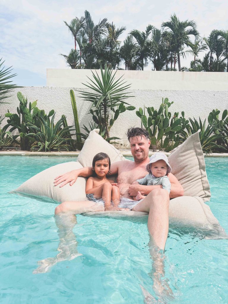A man relaxes on a floating bean bag in a pool with two young children, surrounded by tropical plants and palm trees under a partly cloudy sky near Seseh Beach.