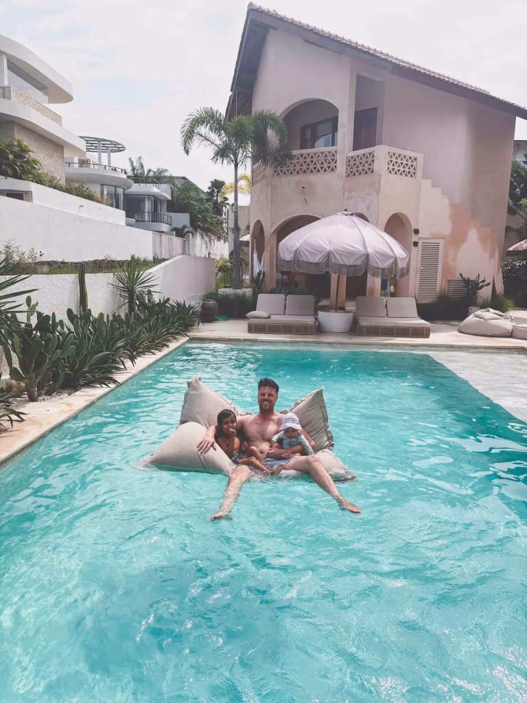A man and a young child relax on a floating cushion in a clear blue pool, with a villa, greenery, and poolside loungers shaded by a large umbrella in the background.