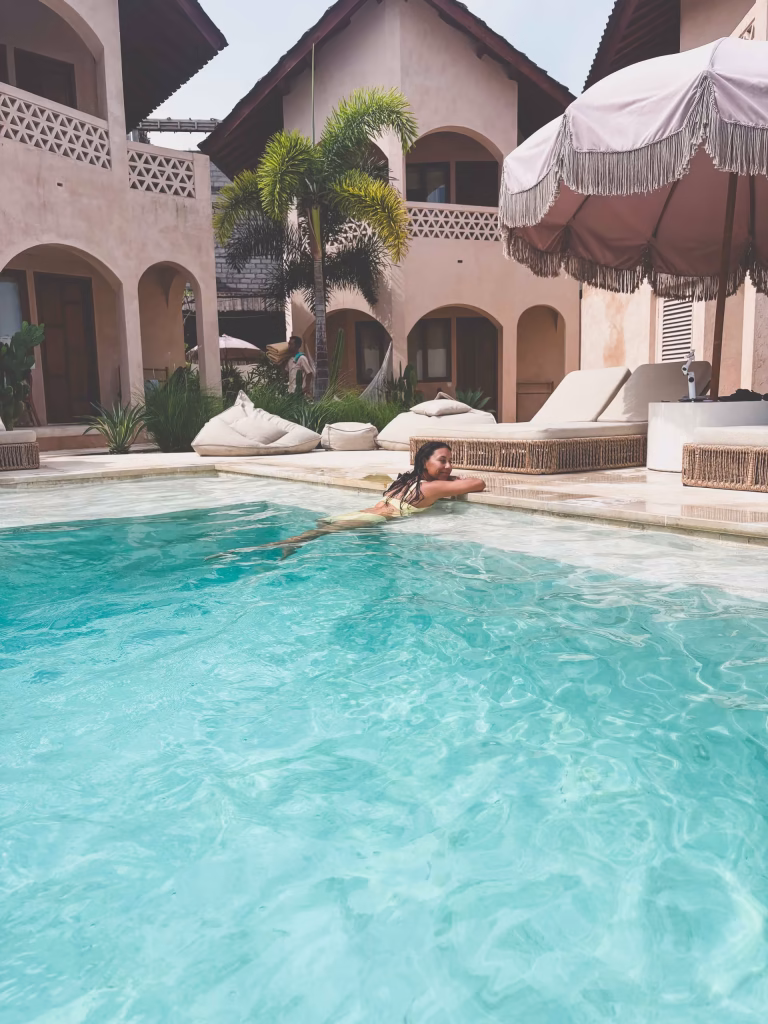 A woman swims in a clear turquoise pool at a resort with light stucco buildings, arched doorways, palm trees, lounge chairs, and a large umbrella in the background. The scene is bright and relaxed.