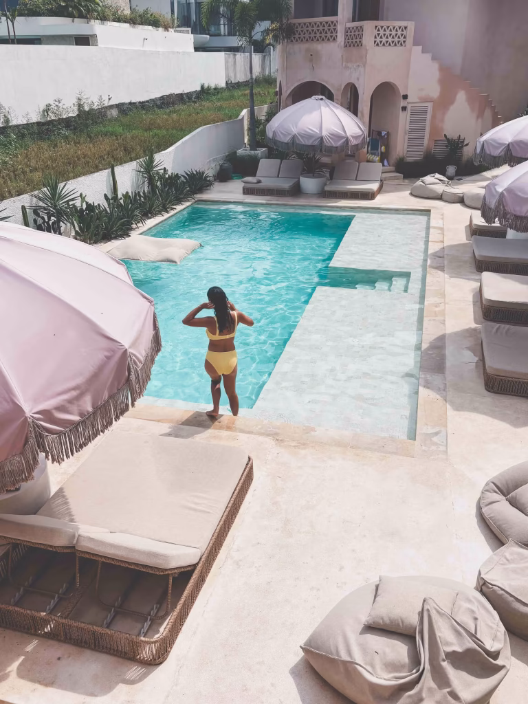A woman in a yellow swimsuit stands at the edge of a swimming pool surrounded by beige lounge chairs and large pink umbrellas on a sunny day. The setting appears to be a modern, stylish outdoor pool area.