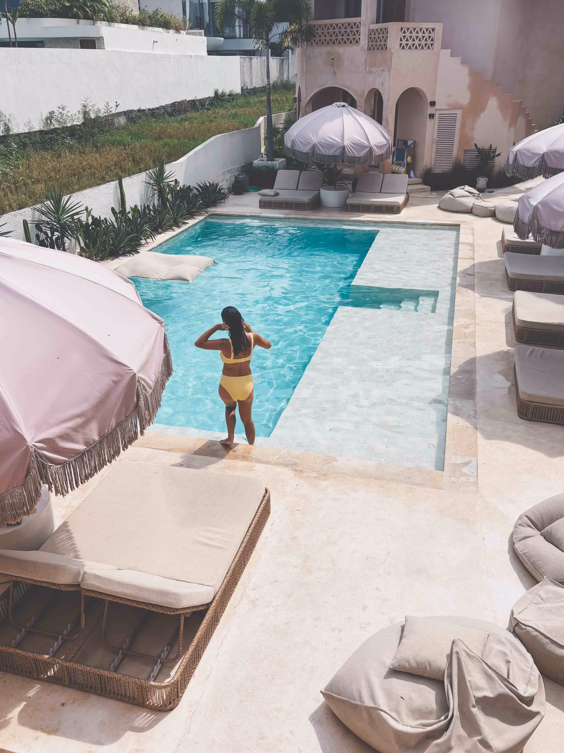 A woman in a yellow swimsuit stands at the edge of a swimming pool surrounded by beige lounge chairs and large pink umbrellas on a sunny day. The setting appears to be a modern, stylish outdoor pool area.
