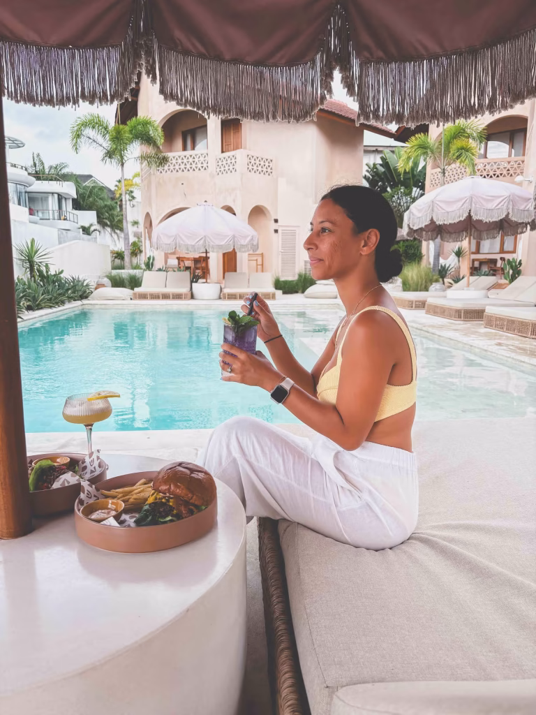 A woman in a yellow bikini top and white pants sits on a poolside lounge bed, holding a drink and smiling. There’s food and another drink on a table in front of her, with a sunny resort and pool in the background.