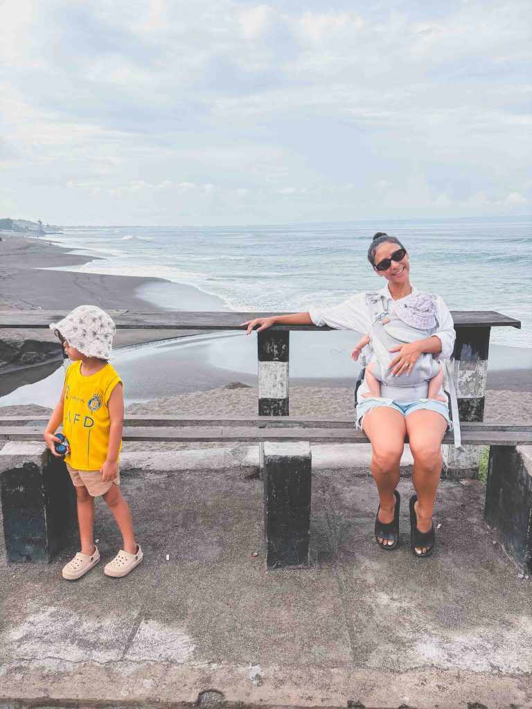A woman sits on a beachside fence at Seseh Beach, holding a baby in her arms. Next to her, a small child in a yellow shirt and sunhat stands looking away. The ocean and cloudy sky form the backdrop.