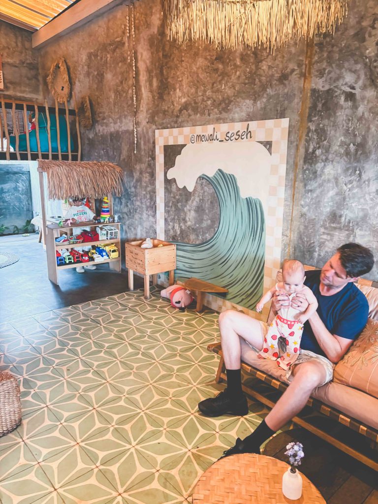 A man sits on a couch holding a baby inside a cozy, rustic playroom inspired by Seseh Beach, with a wave mural, patterned tile floor, toy-filled shelves, high ceiling, and a small loft area.