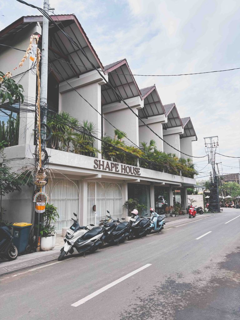 A row of modern buildings with balconies and plants, labeled Shape House, lines a street near Seseh Beach, where parked scooters and motorcycles rest under a cloudy sky.