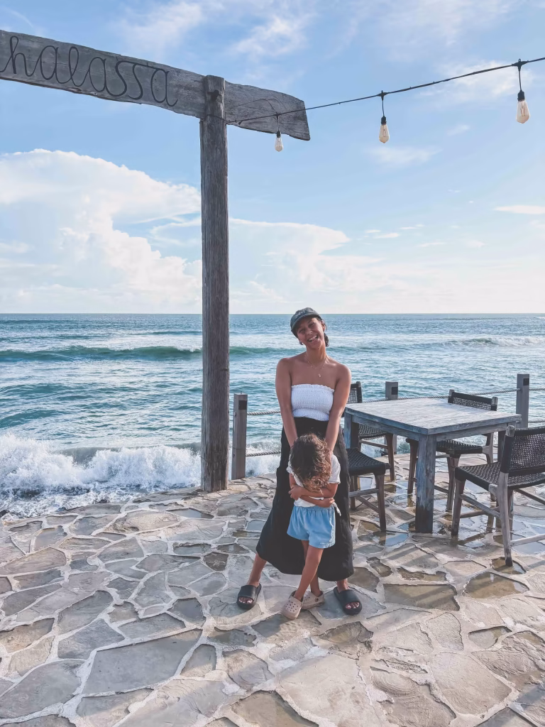 A woman and a child stand smiling on a stone patio by the ocean, with waves in the background. Wooden tables and chairs are nearby, and a rustic wooden sign hangs above them under string lights.