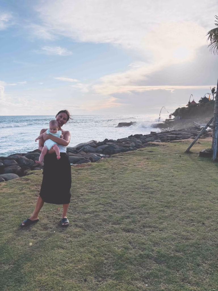 A woman stands on a grassy area near rocky shoreline, holding a baby. Waves crash against the rocks behind them under a cloudy sky, with the sun low on the horizon.