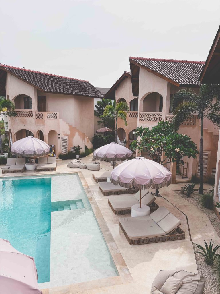 A serene courtyard with a turquoise swimming pool, surrounded by beige loungers and large white umbrellas. Two-story villas with tiled roofs and palm trees border the peaceful area, just moments from Seseh Beach.
