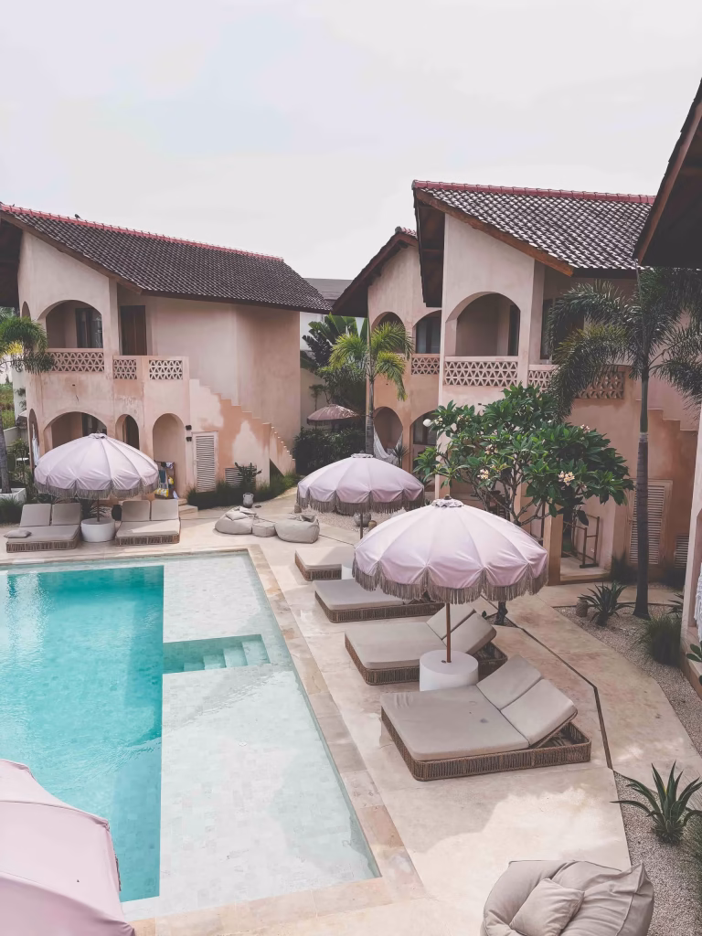 A luxurious outdoor pool area features white lounge chairs with large pale pink umbrellas, surrounded by beige Mediterranean-style buildings with balconies, palm trees, and greenery. The setting looks serene and inviting.
