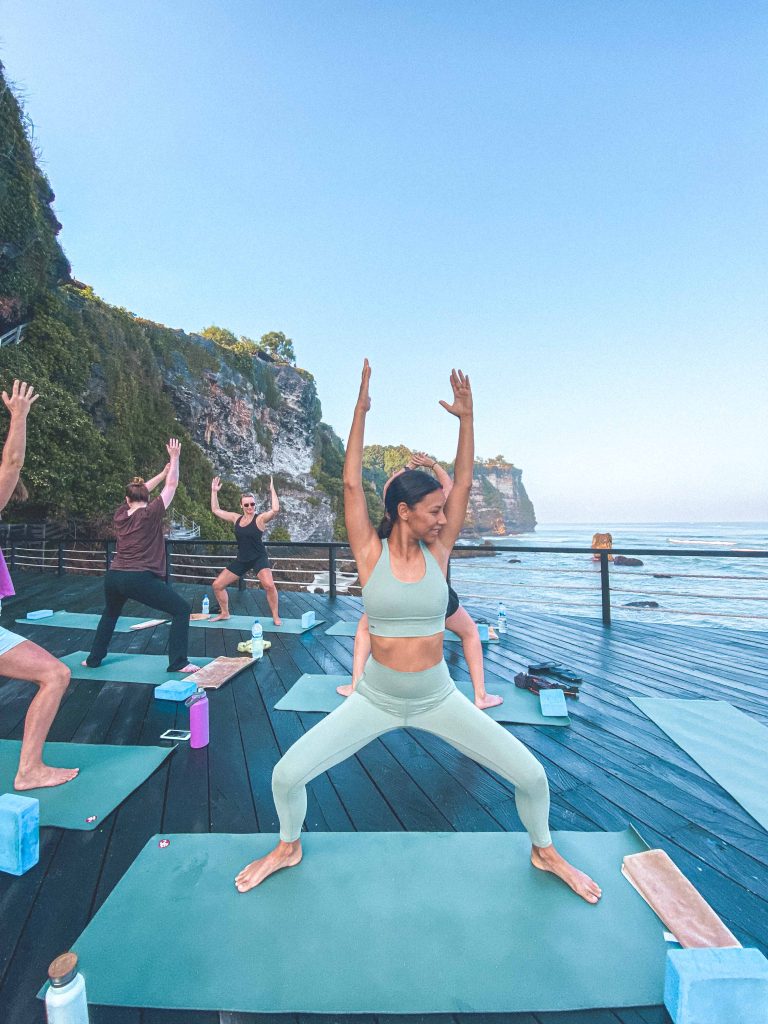 A group practices yoga on mats outdoors at Uluwatu Surf Villas, on a wooden deck by the ocean and cliffs. One woman in light blue activewear raises her arms, smiling in the clear, sunny weather—perfect for a Bali wedding retreat.