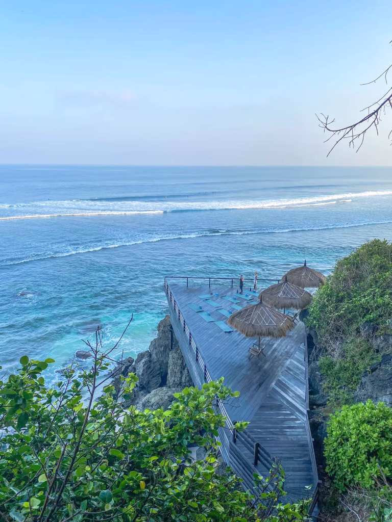 A wooden deck with straw umbrellas extends over rocky cliffs above turquoise ocean waves, surrounded by green foliage, under a clear blue sky.