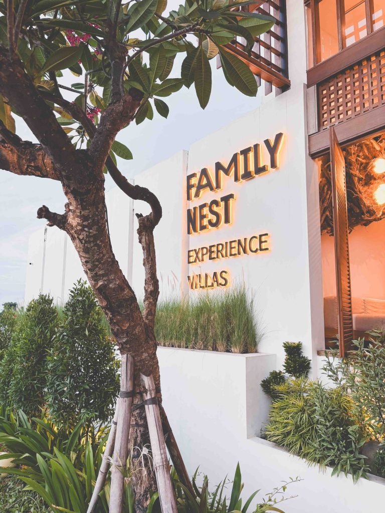 Family Nest Uluwatu Villas. A modern white building with illuminated signage reading FAMILY NEST EXPERIENCE VILLAS, surrounded by lush green plants and a tree in the foreground.