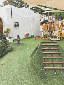 Pangolin Kids Club outside playground. A child stands near a trampoline and basketball hoop in a playground with artificial grass, a small climbing hill with a rope ladder, colorful toys, and a large play structure in the background.